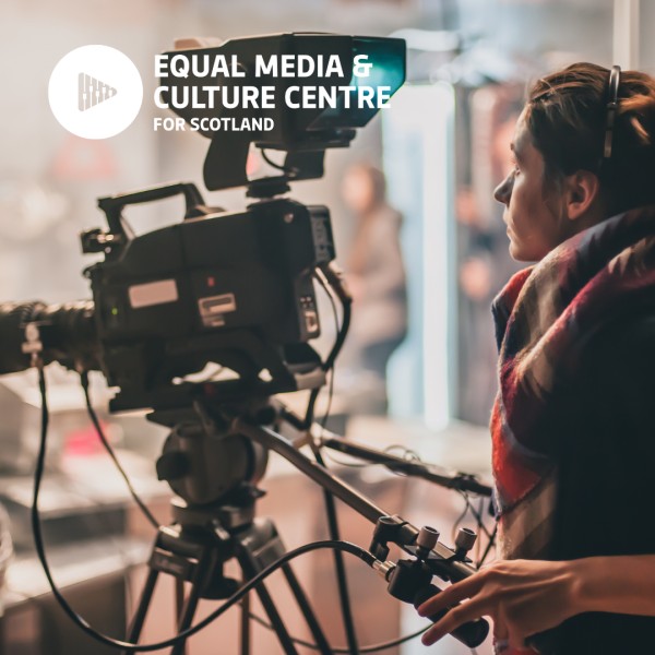 A photograph of a white woman with her hair in a bun operating a camera on a film set