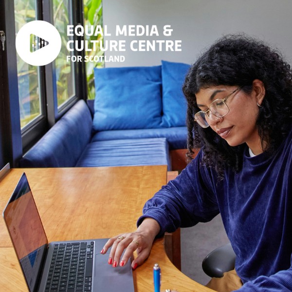A photograph of a woman with brown skin and black curly hair working at a laptop on a wooden desk. She wears glasses and a blue jumper. She sits by a window. 