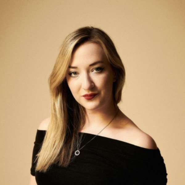 a photograph of singer Iona Fyfe, a white woman with long blonde hair. She wears a black top and is photographed in front of a cream background.